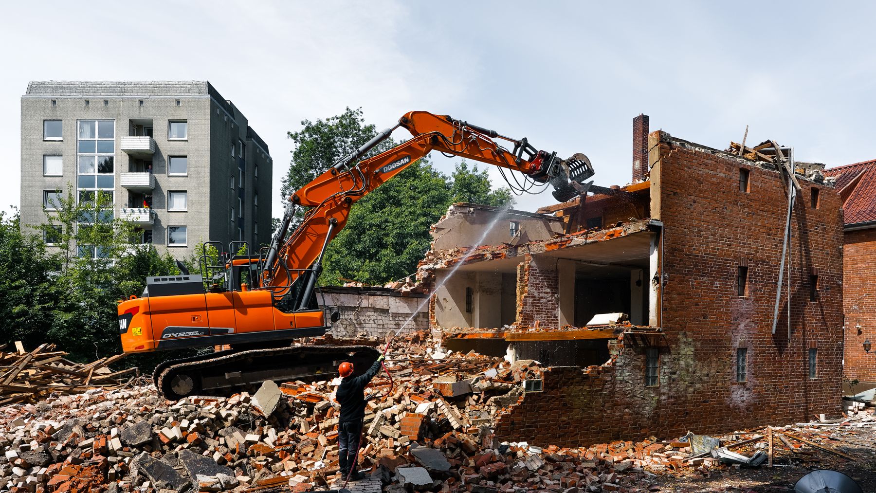 An orange excavator with a grapple pulling apart a brick building while a worker in a hard hat sprays water on the rubble to keep dust down.